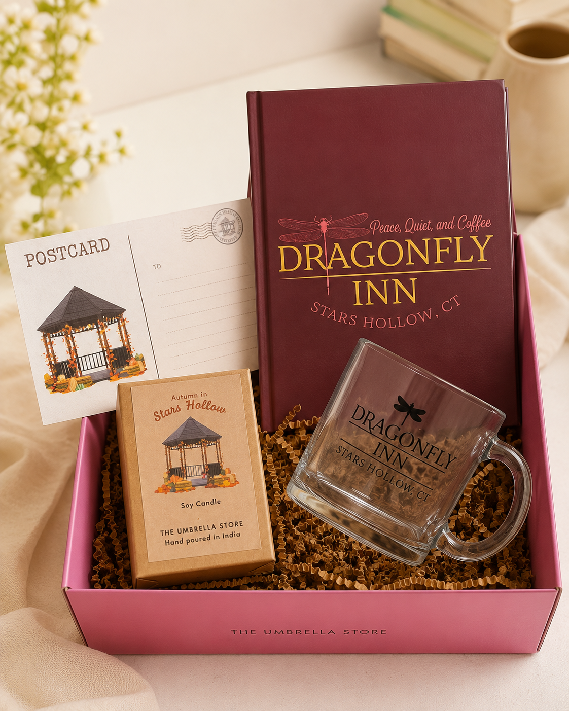 A Dragonfly Inn themed gift box set from The Umbrella Store, featuring a deep burgundy tumbler, a clear glass mug, a small 'Star Candle' box, a postcard, and a letter opener, all arranged in a pink box with brown crinkle paper filler.