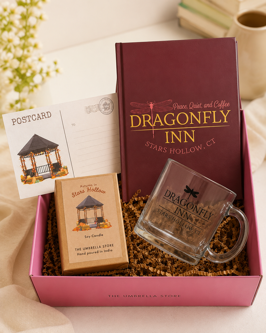 A Dragonfly Inn themed gift box set from The Umbrella Store, featuring a deep burgundy tumbler, a clear glass mug, a small 'Star Candle' box, a postcard, and a letter opener, all arranged in a pink box with brown crinkle paper filler.